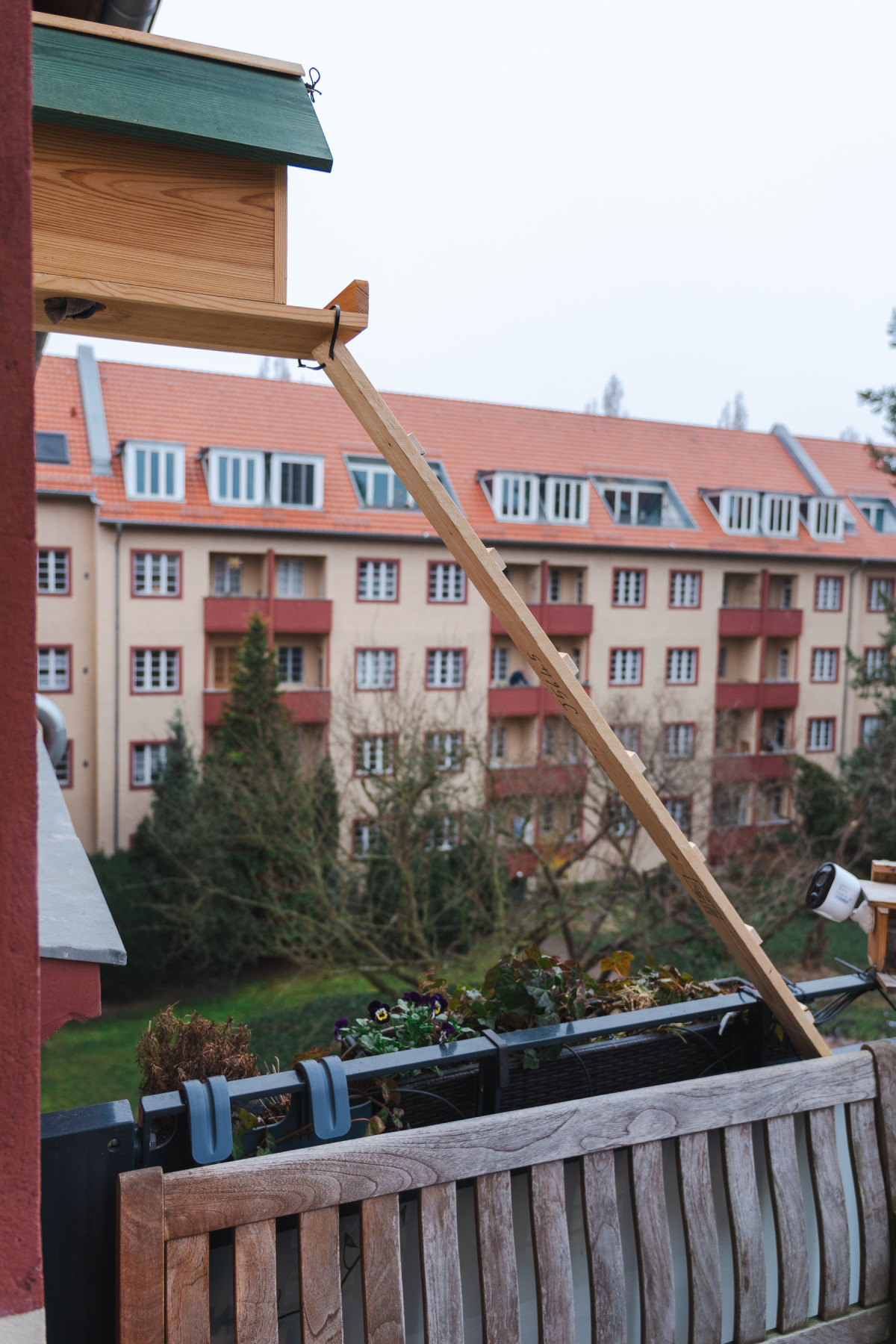 Der Kobel hängt an der Hauswand. Darunter ist der Balkon mit Blumenkästen. Von den Blumenkästen aus führt nun eine  Art Hühnerleiter aus Holz hoch zum Kobel.